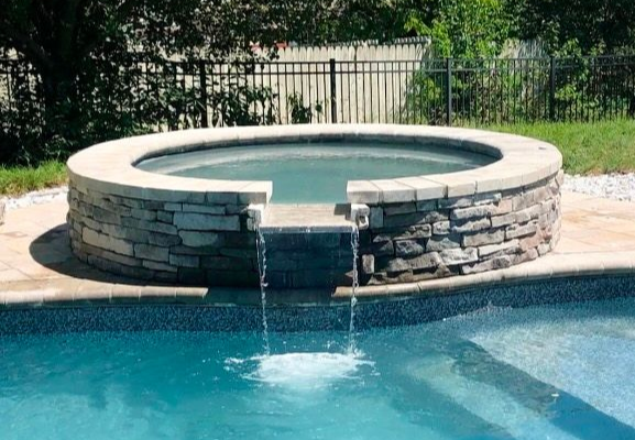 Circular stone hot tub spilling water into a turquoise swimming pool in a backyard.
