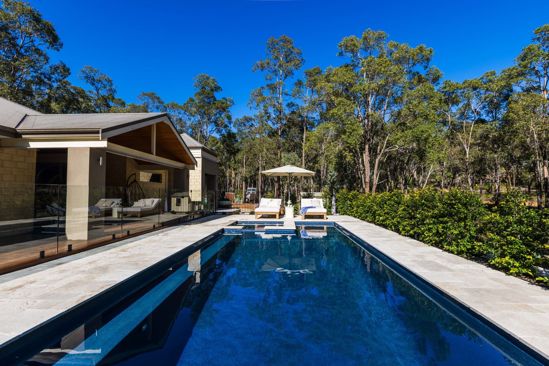 Long backyard pool beside a modern house, with lounge chairs and trees under a clear blue sky