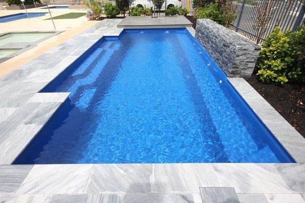 Rectangular blue swimming pool with light stone decking and landscaping around it