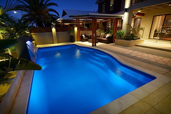 Illuminated backyard pool at night with blue water, patio lights, and tropical plants
