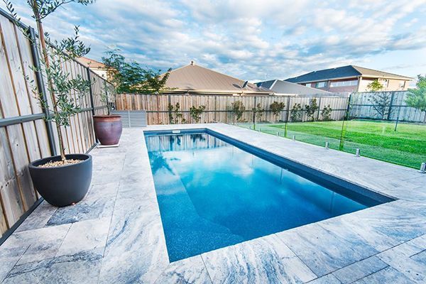 Rectangular backyard swimming pool with blue water, surrounded by stone patio and fenced lawn.