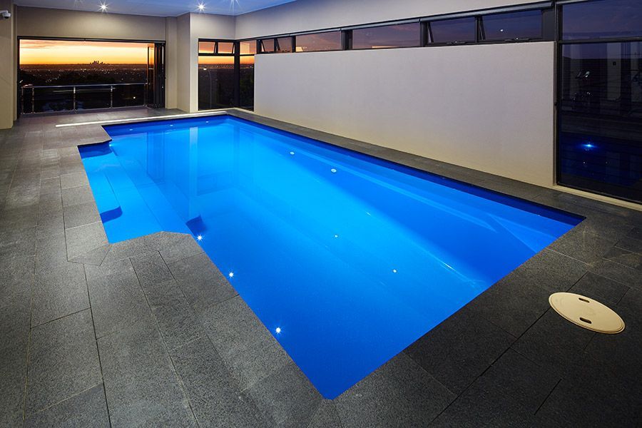 Indoor pool with glowing blue water beside a tiled deck at dusk