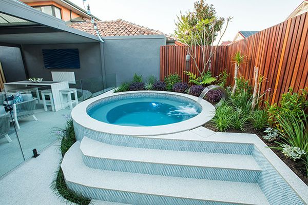 Small backyard spa pool with white steps, blue water, and a modern patio beside greenery and a wooden fence