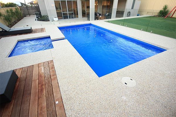 Modern backyard with a blue swimming pool, spa, and wooden deck beside a patio.