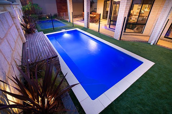 Backyard with a lit blue rectangular pool, stone seating area, and patio beside a house at dusk
