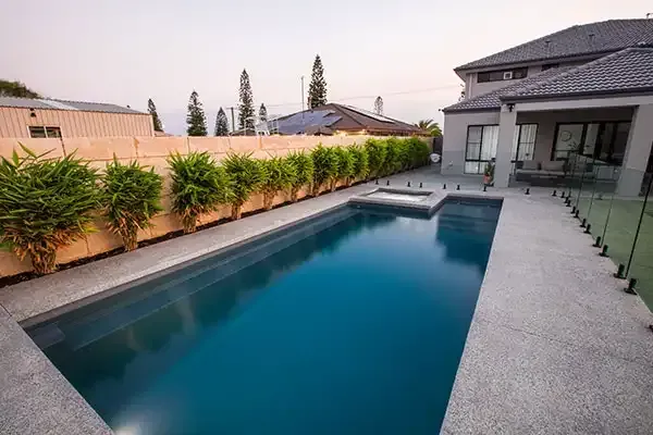 Backyard swimming pool beside a modern house at dusk, with hedge-lined fence and lounge chairs