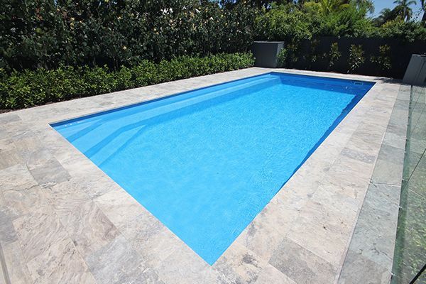 Rectangular backyard swimming pool with bright blue water, bordered by light stone and greenery