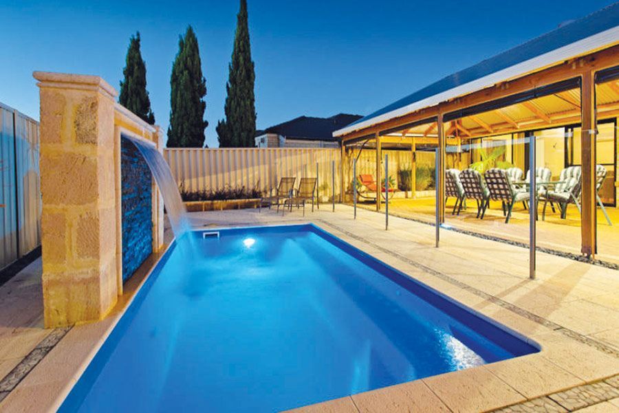 Backyard pool with a water feature, lit at dusk beside a covered patio and lounge area