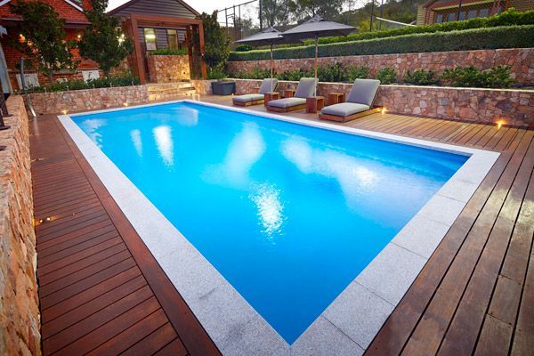 Outdoor backyard pool with bright blue water, wooden deck, lounge chairs, and warm garden lighting