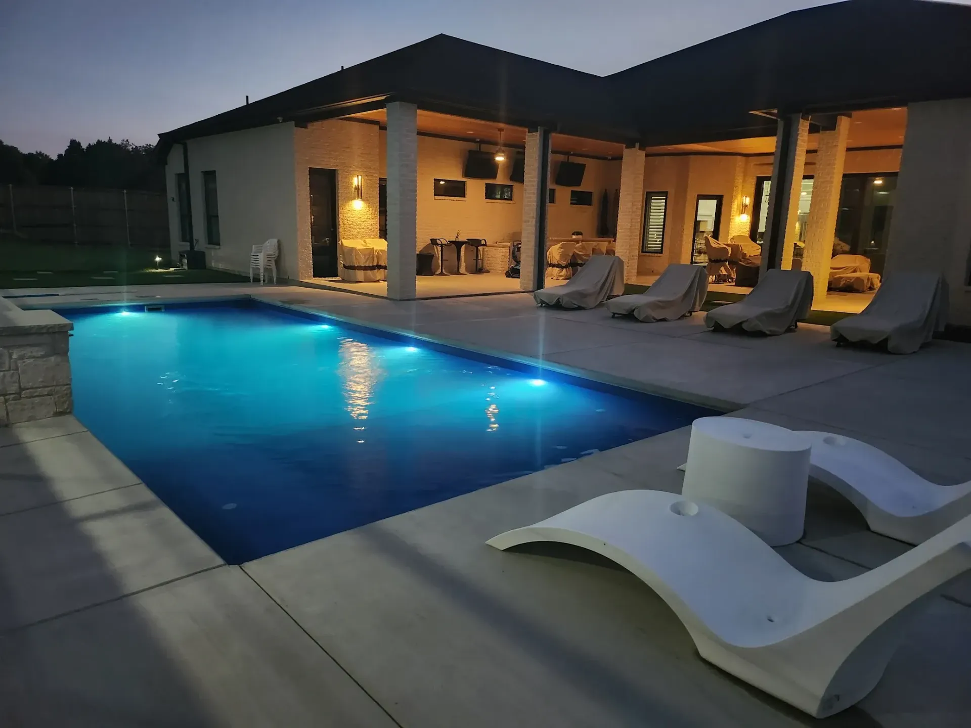 Modern house patio with glowing blue swimming pool at dusk and lounge chairs under warm lights