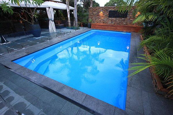 Small backyard swimming pool with bright blue water, surrounded by stone paving and lush greenery.