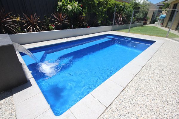 Small backyard swimming pool with bright blue water and a fountain spray, surrounded by patio and plants