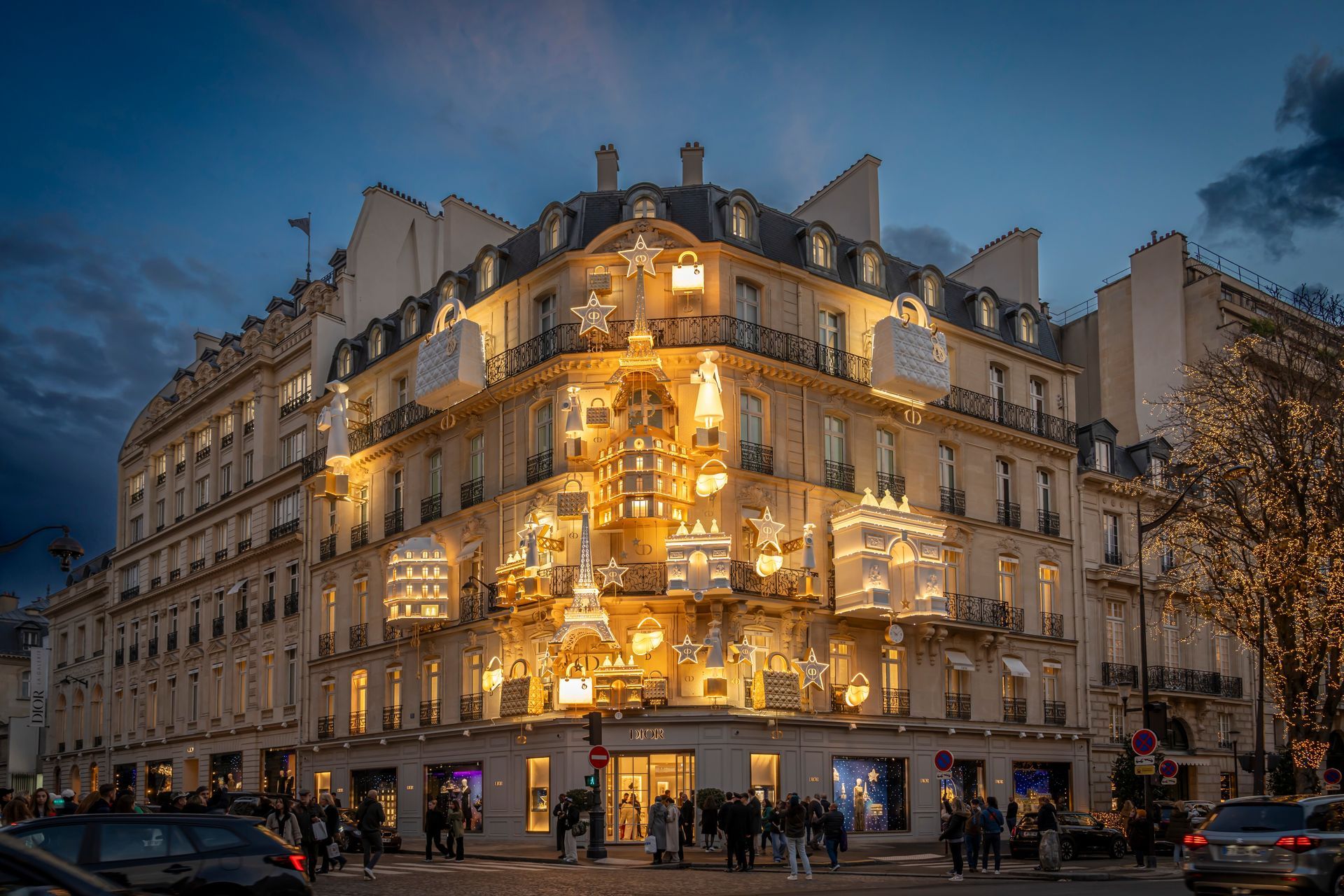 Illuminated luxury building in Paris at dusk. Corner storefront with multiple lit levels. People on the sidewalk.