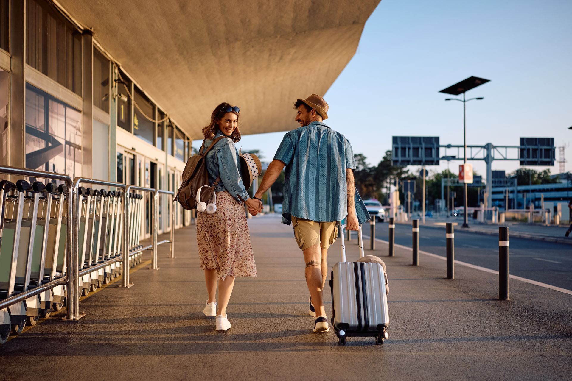 Couple walking, holding hands, near a building. One pulls a suitcase. Sunny, outdoor setting.