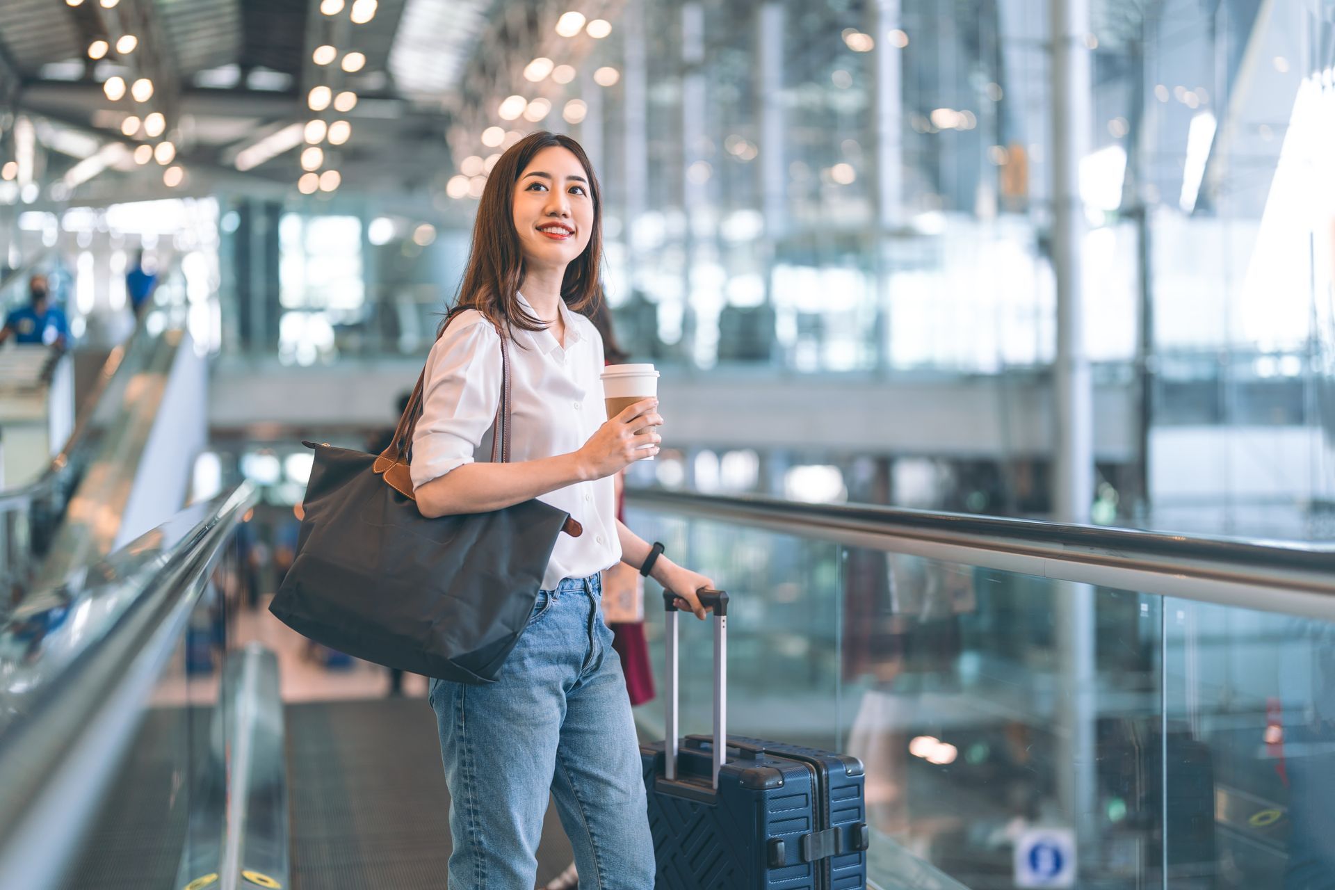 Woman in an airport, smiling, holding coffee and a bag, pulling a suitcase.