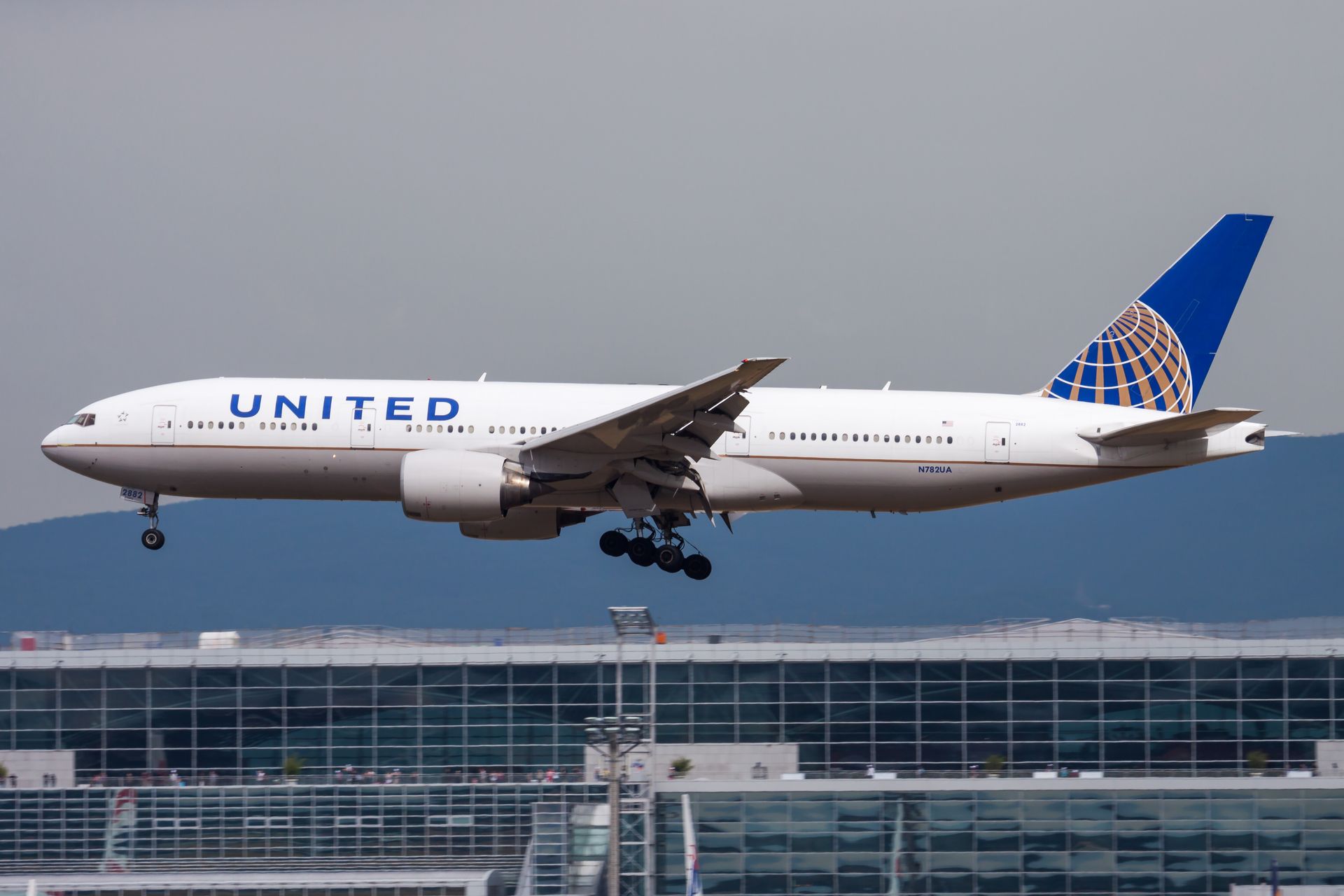 United Airlines Boeing 777 approaching for landing, over a modern building with blue and white accents.