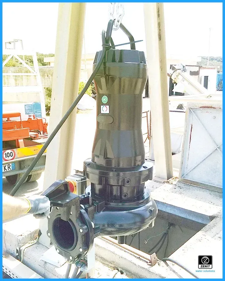 Pompa sommergibile calata in un pozzo, nera e argento, appesa a una catena, all'aperto.