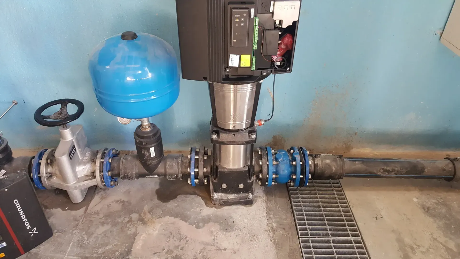 Sistema di pompaggio dell'acqua con serbatoio di pressione blu, valvola argentata e tubature nere contro una parete blu.