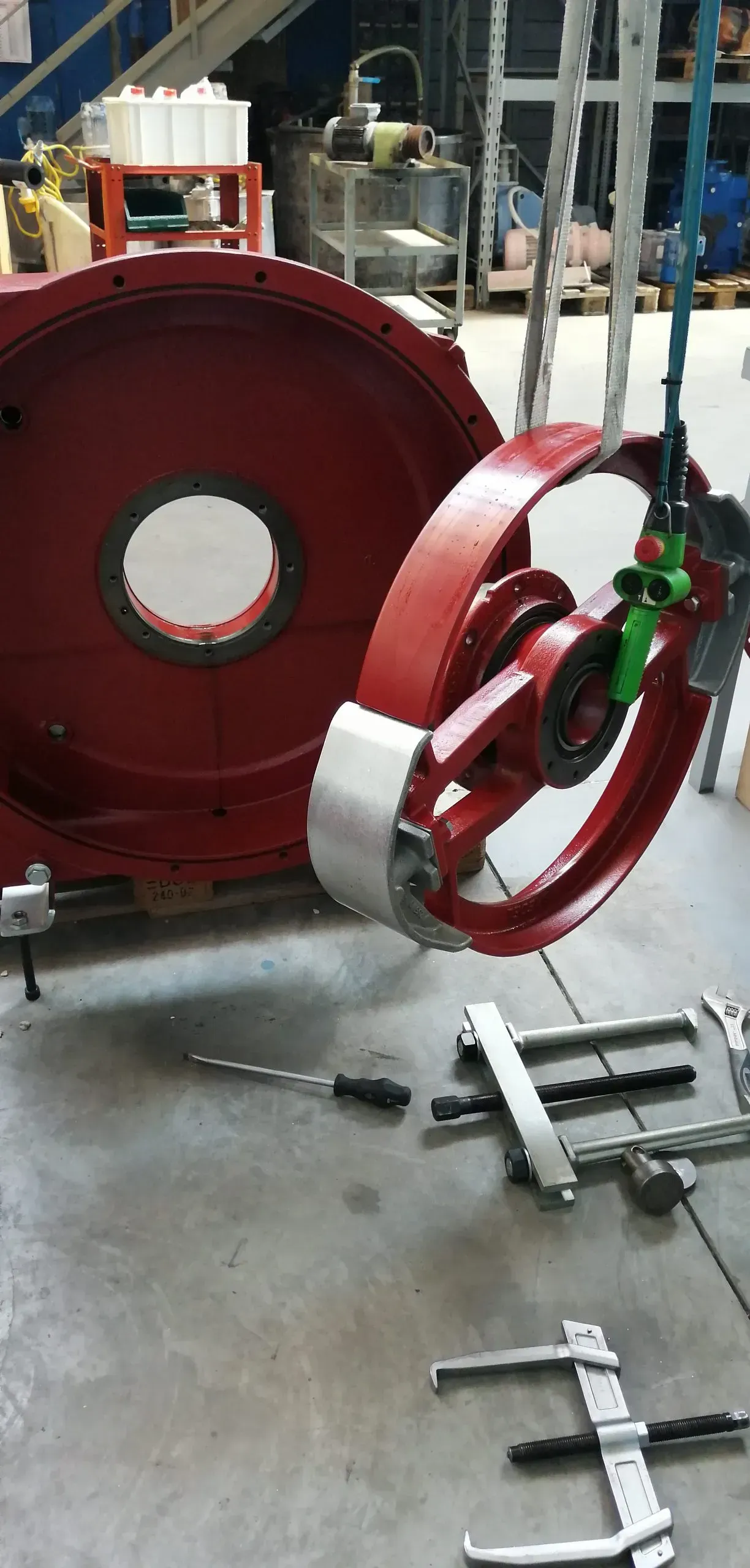 Un grande componente industriale rosso viene sollevato da una gru, con degli attrezzi e un pezzo rosso più grande sul pavimento.