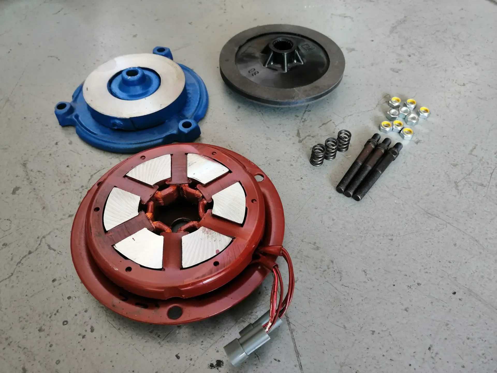 Parti smontate della frizione elettromagnetica in blu, nero e rosso; sono inclusi bulloni e un connettore per cavi.