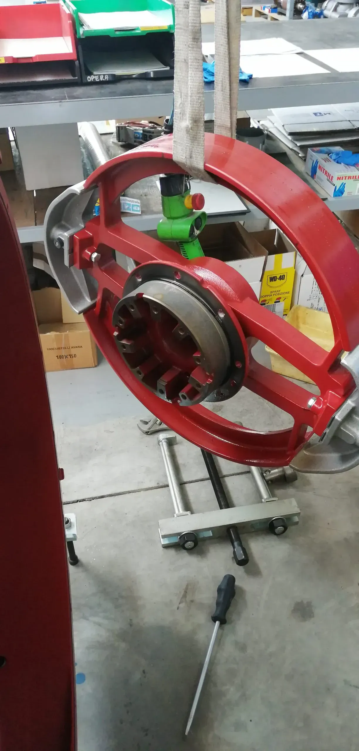 Un grande componente rosso di una macchina è sospeso su un supporto metallico, con un cacciavite nelle vicinanze.