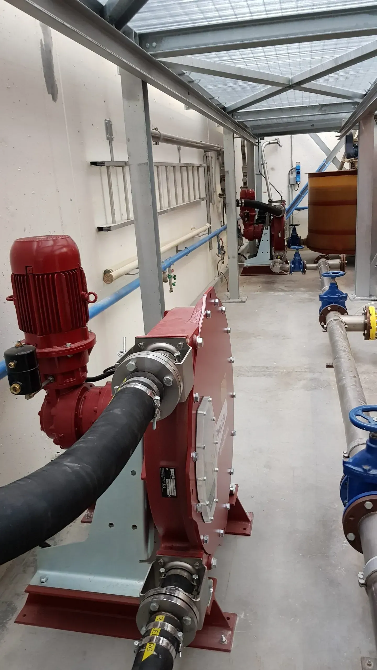 Impianto industriale con pompe, tubi e serbatoi rossi e argento in un lungo corridoio sotto un soffitto a griglia metallica.