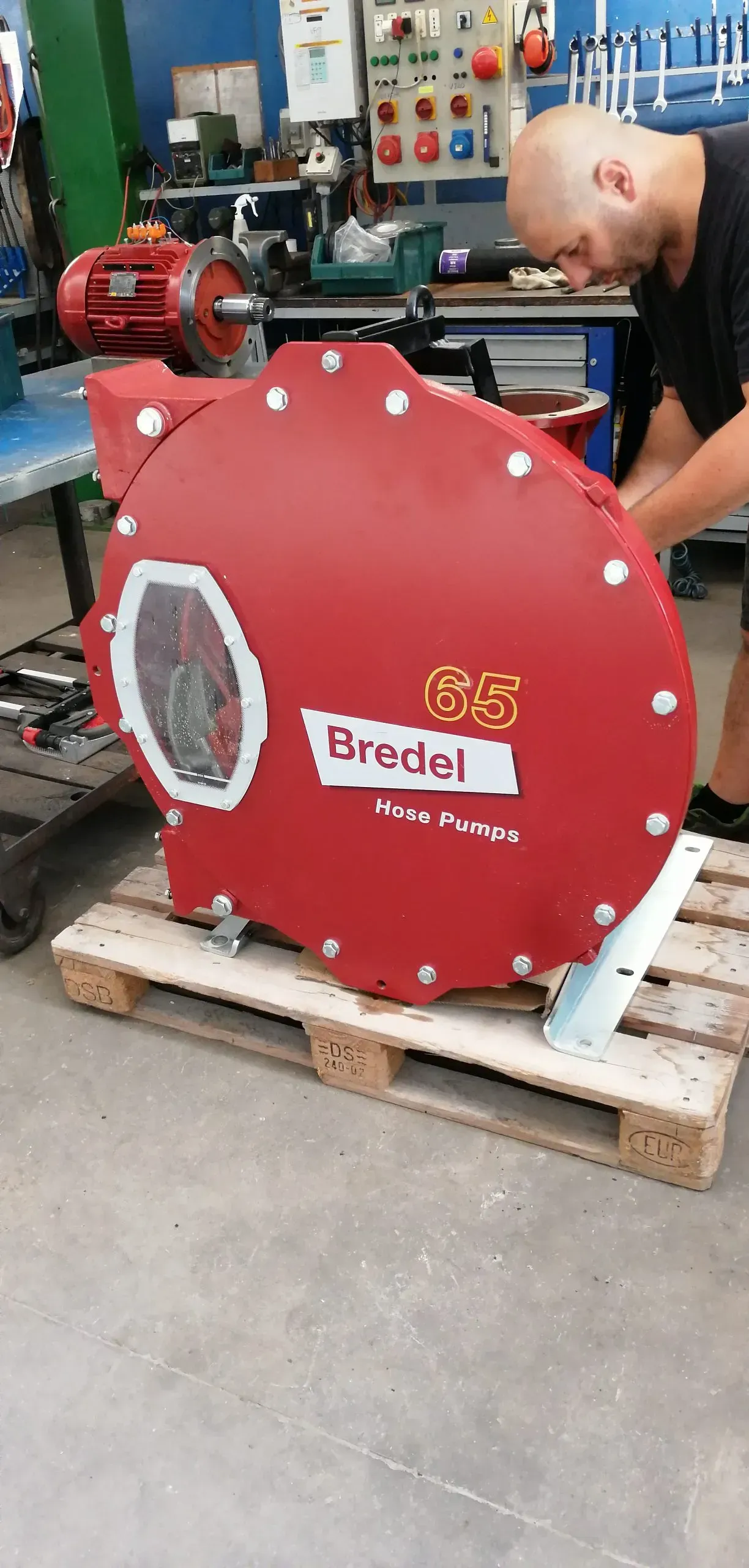 Una persona lavora su una grande pompa rossa Bredel appoggiata su un pallet in un'officina.