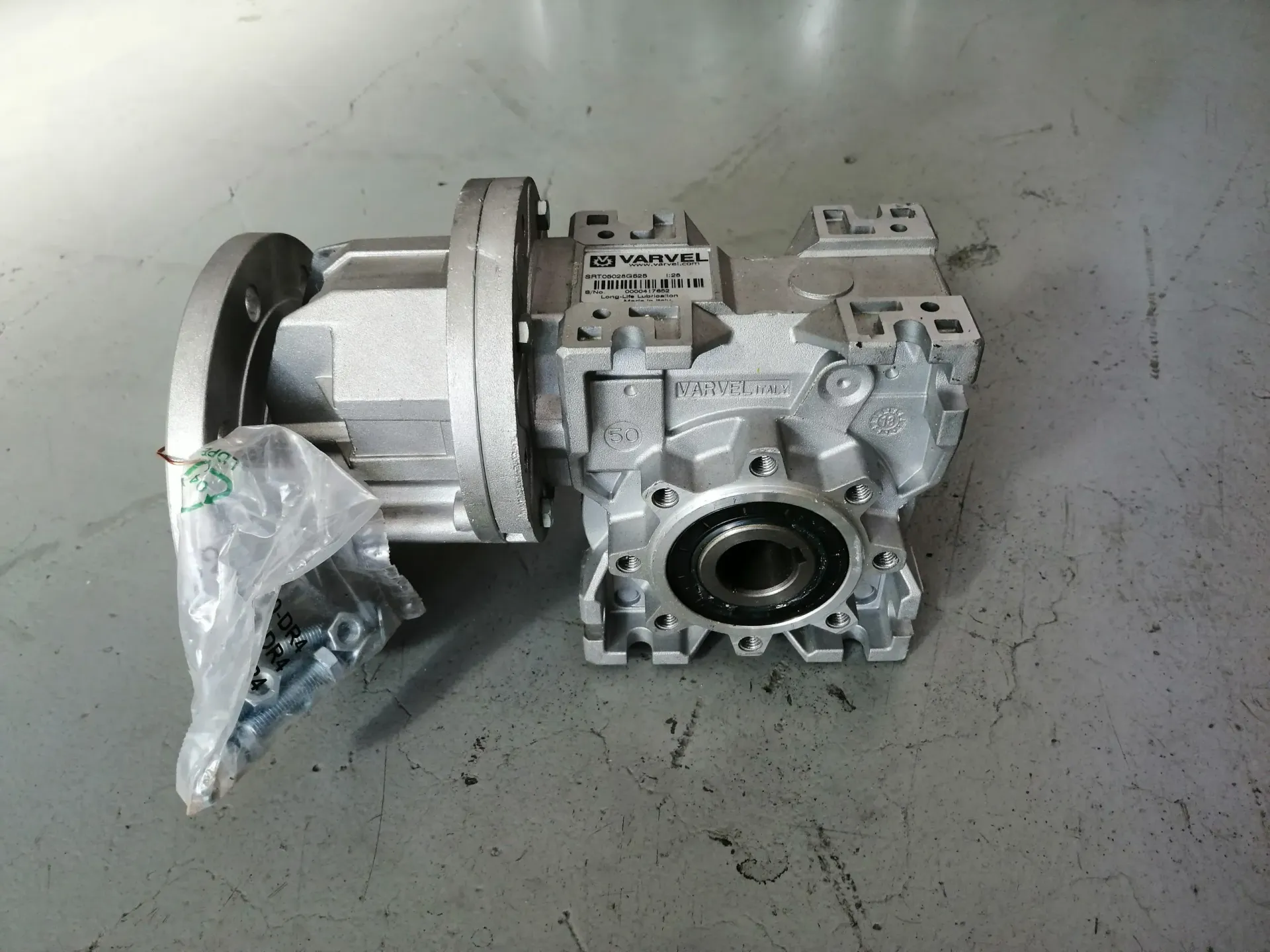 Scatola del cambio argentata con flangia, pellicola di plastica e caratteristiche di montaggio, su una superficie grigia.