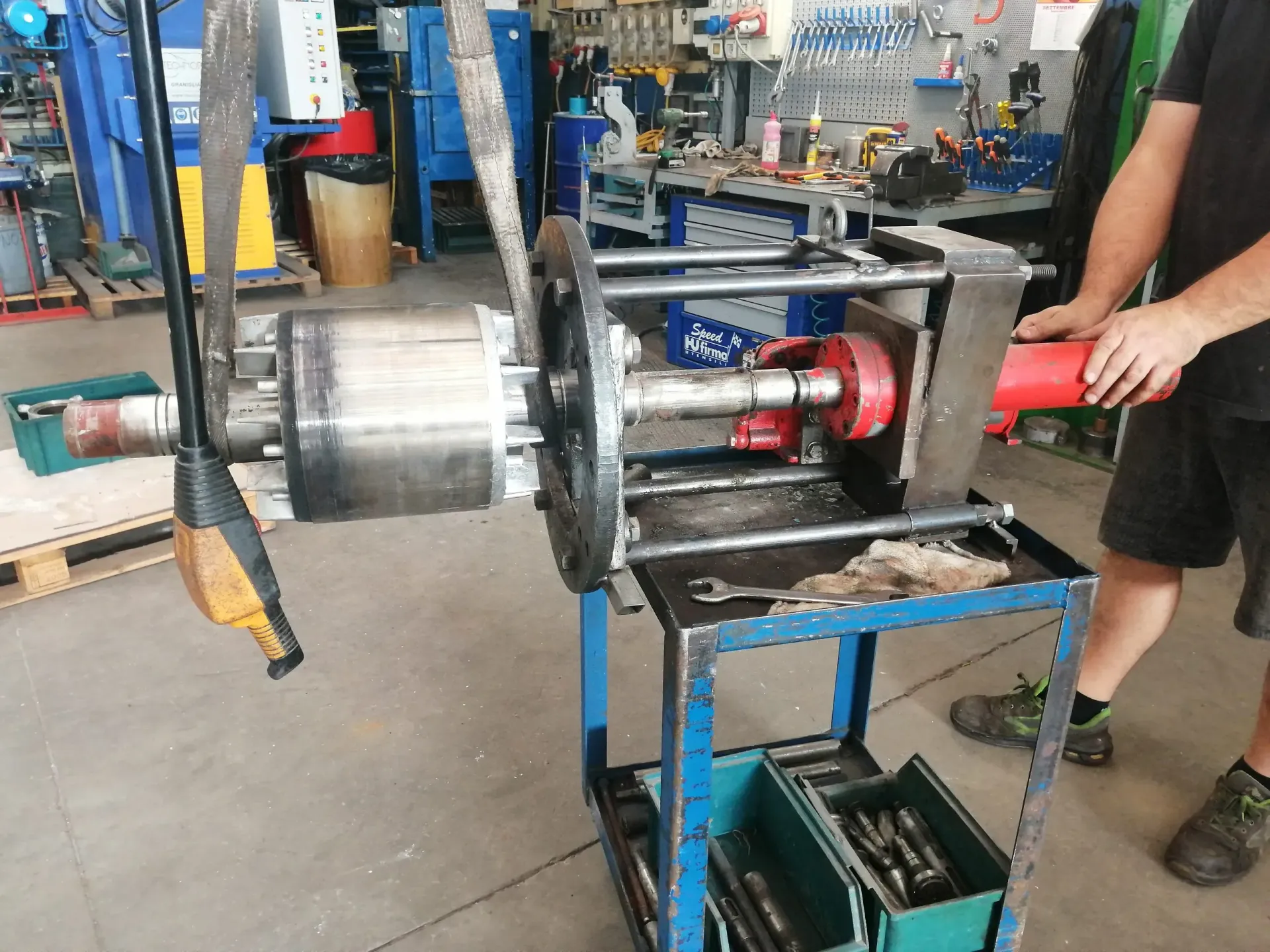 Persona che lavora su macchinari industriali in un'officina. Un grande cilindro metallico è fissato su un carrello.