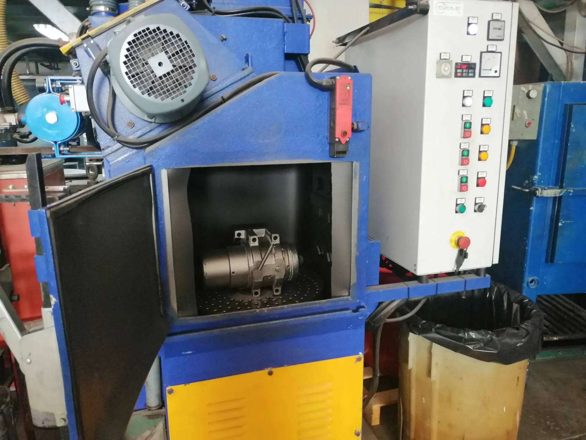 Macchina industriale blu con sportello aperto, contenente parti metalliche. Quadro elettrico e cestino dei rifiuti nelle vicinanze.