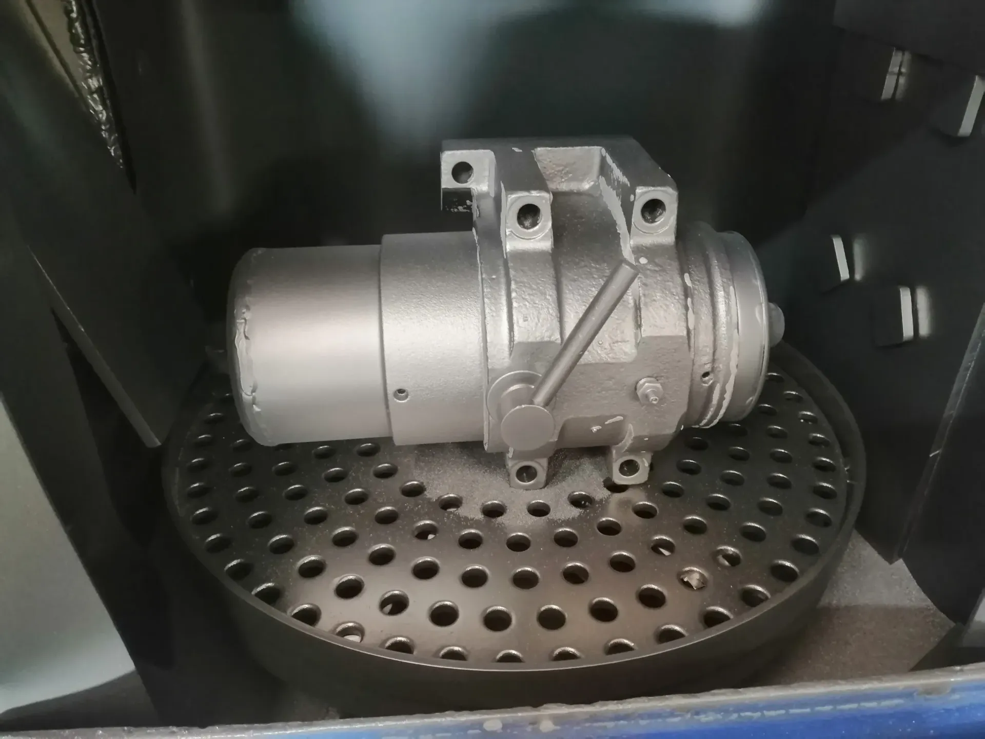 La parte metallica industriale è appoggiata su un vassoio di metallo perforato all'interno di un involucro scuro.