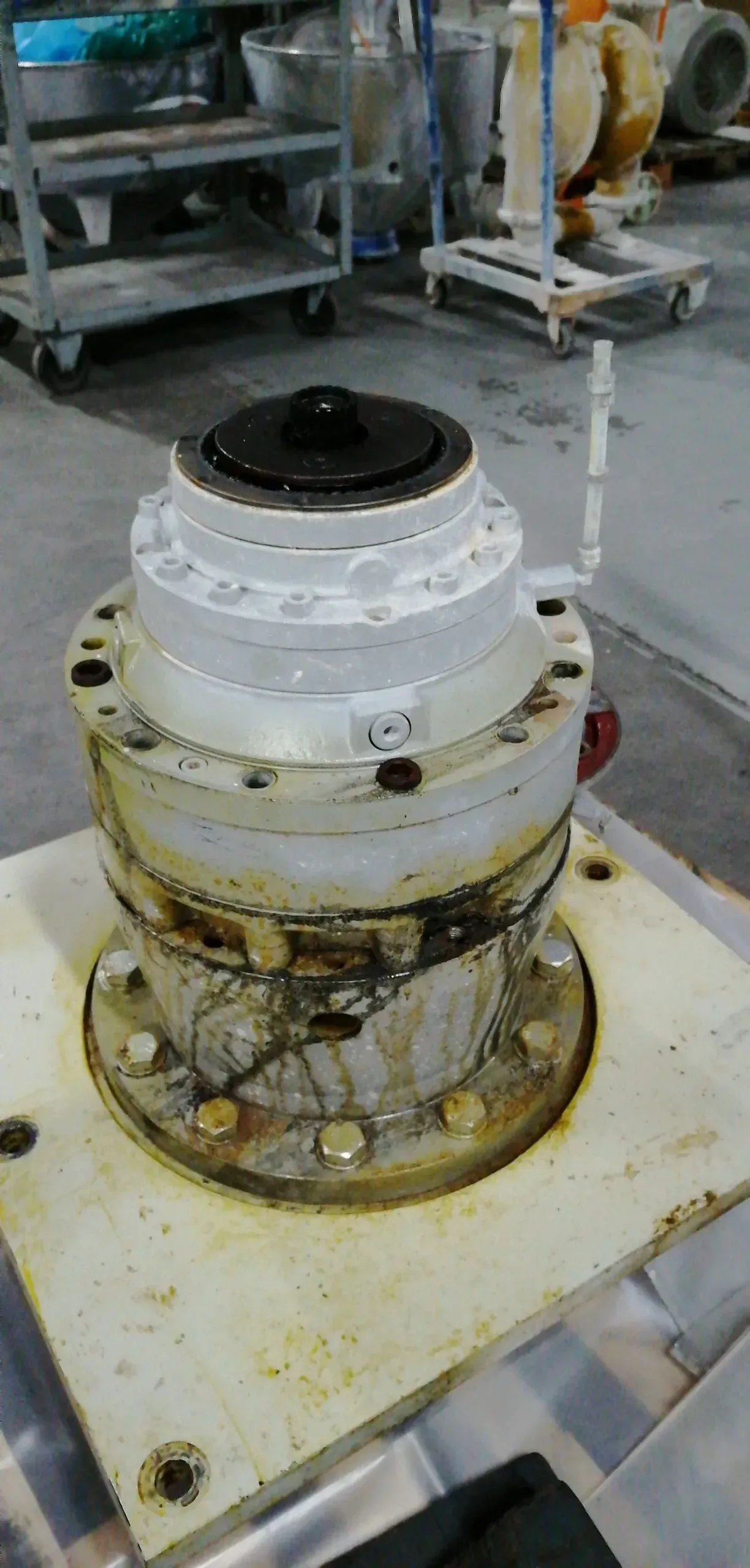 Una macchina industriale in metallo. Toni bianchi e grigi con la parte superiore scura. Sporca per il tempo e l'uso. Fissata a una superficie piana con bulloni.