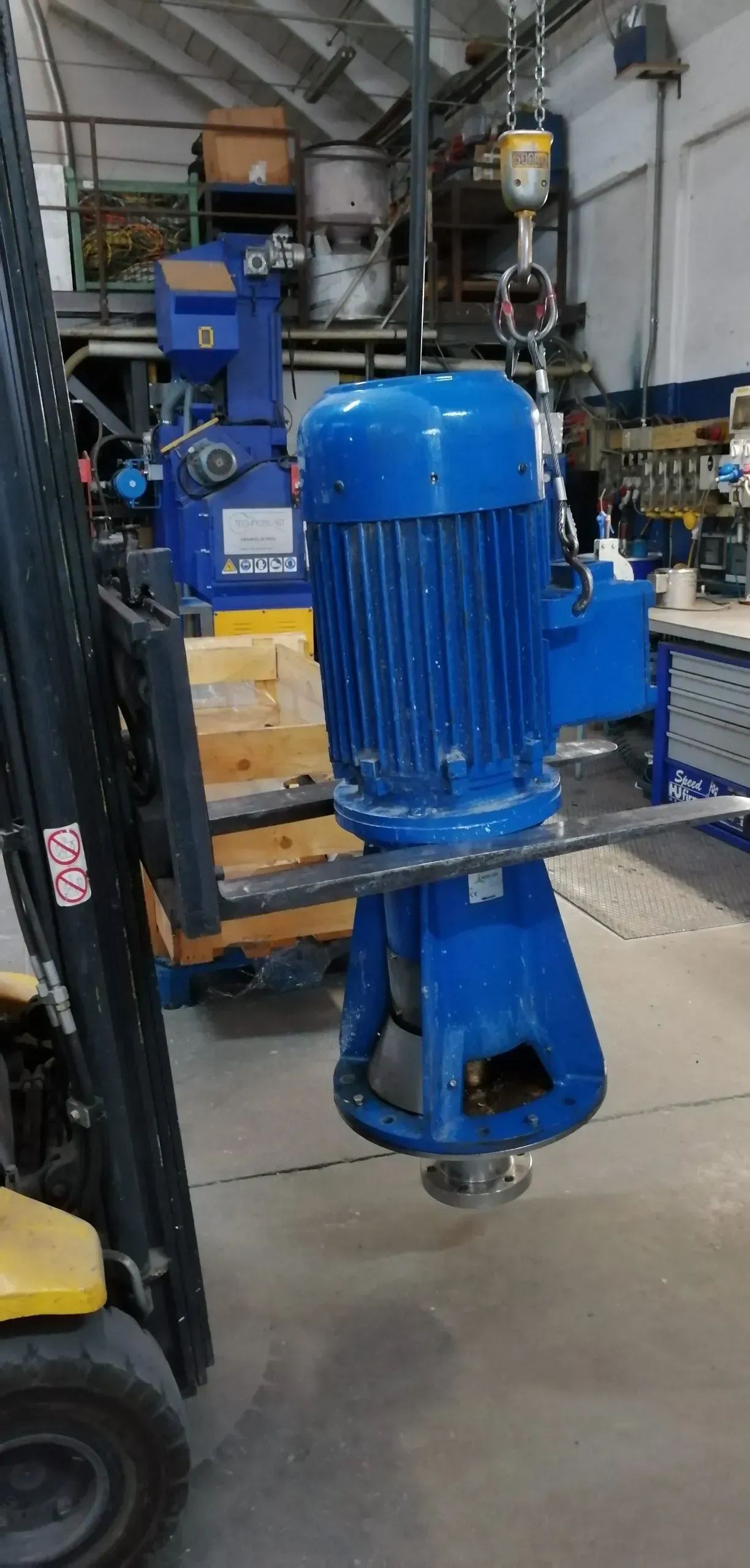 Pompa industriale blu sospesa a una gru in un'officina, vicino a un carrello elevatore.