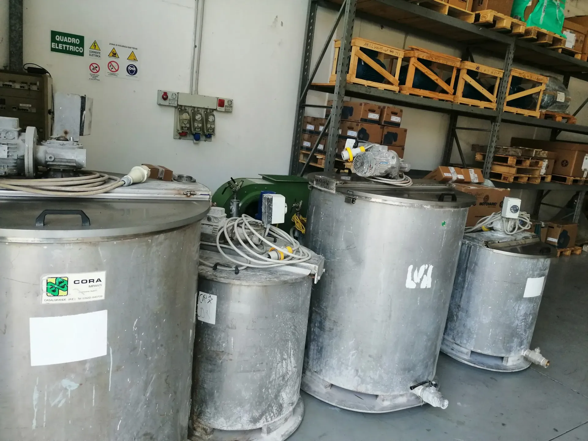 Quattro serbatoi industriali in acciaio inossidabile all'interno di un magazzino, con scaffalature e apparecchiature elettriche sullo sfondo.