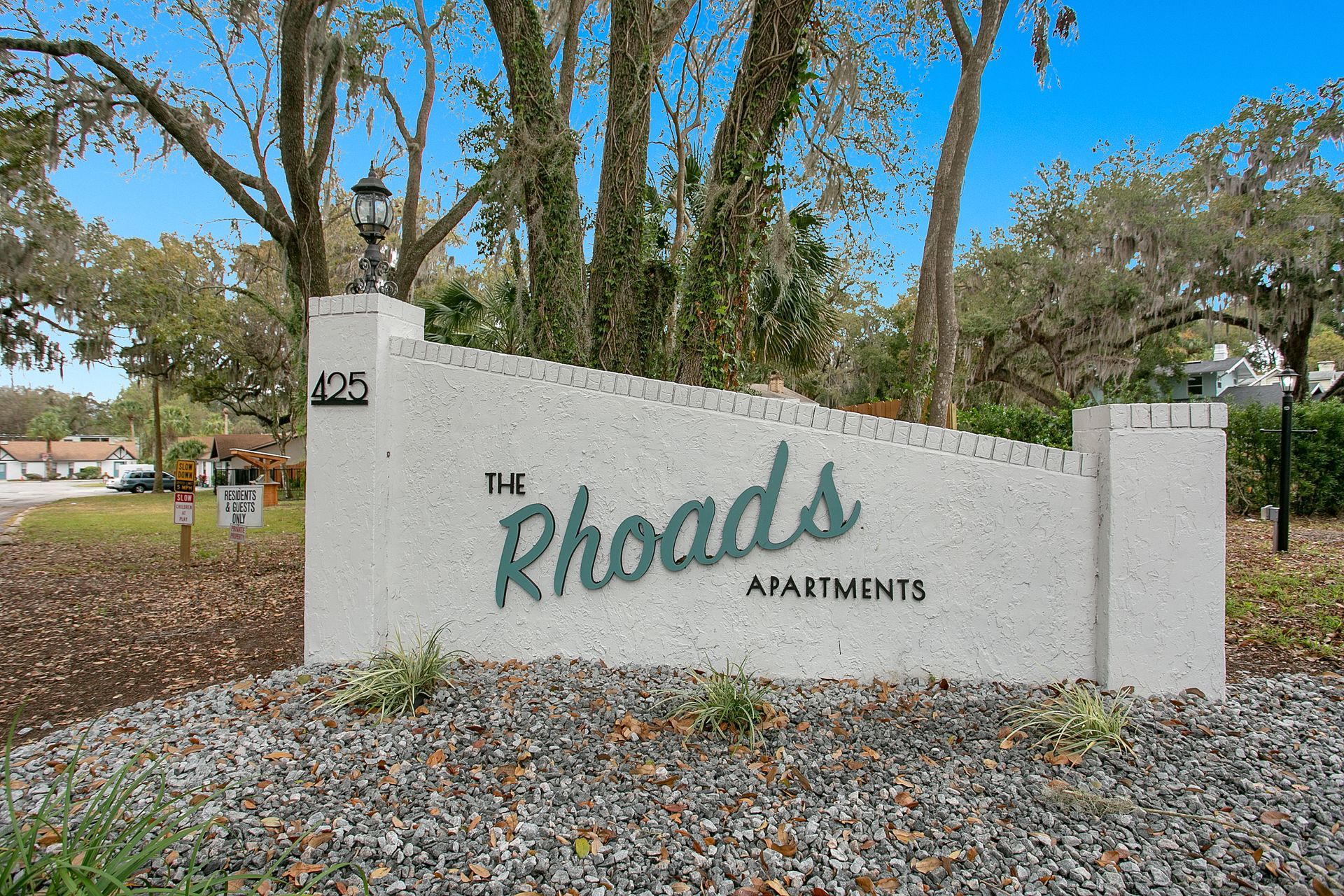 A sign for the rhoads apartments is surrounded by gravel and trees.