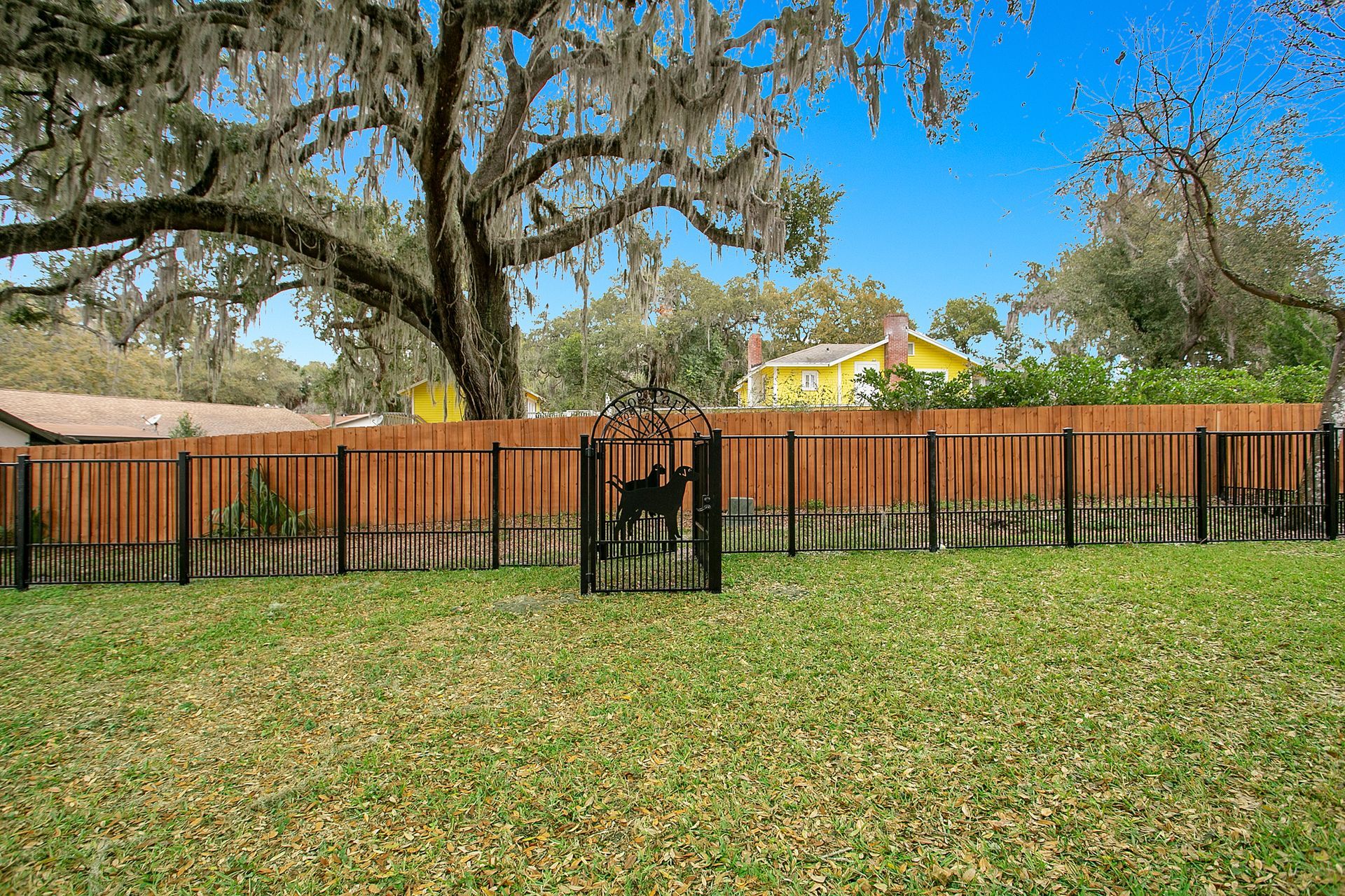 A wooden fence surrounds a lush green yard with a yellow house in the background.