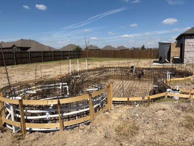 Pool construction site with wooden forms, rebar framework, and plumbing, under a blue sky.
