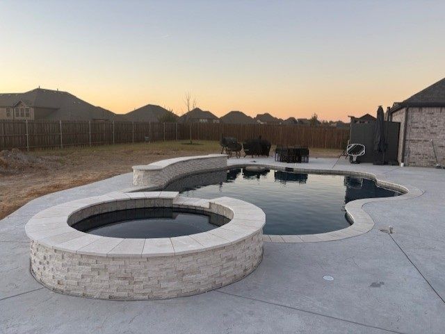 Pool and hot tub with stonework, concrete patio, and seating area at dusk.