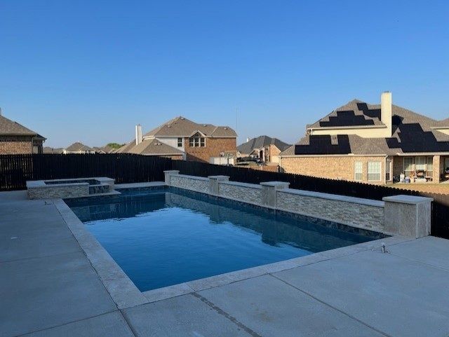 Backyard pool with a dark fence and houses in the background under a blue sky.