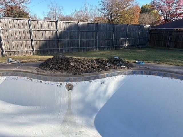 Empty swimming pool with a pile of dirt, set against a wooden fence.