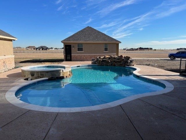 Pool with spa, waterfall, and small building in backyard, blue water under a sunny sky.
