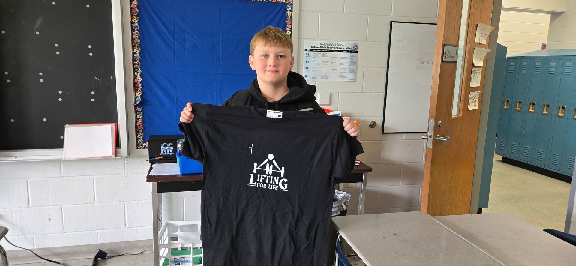 Person holding up a black T-shirt with a white graphic in a classroom setting