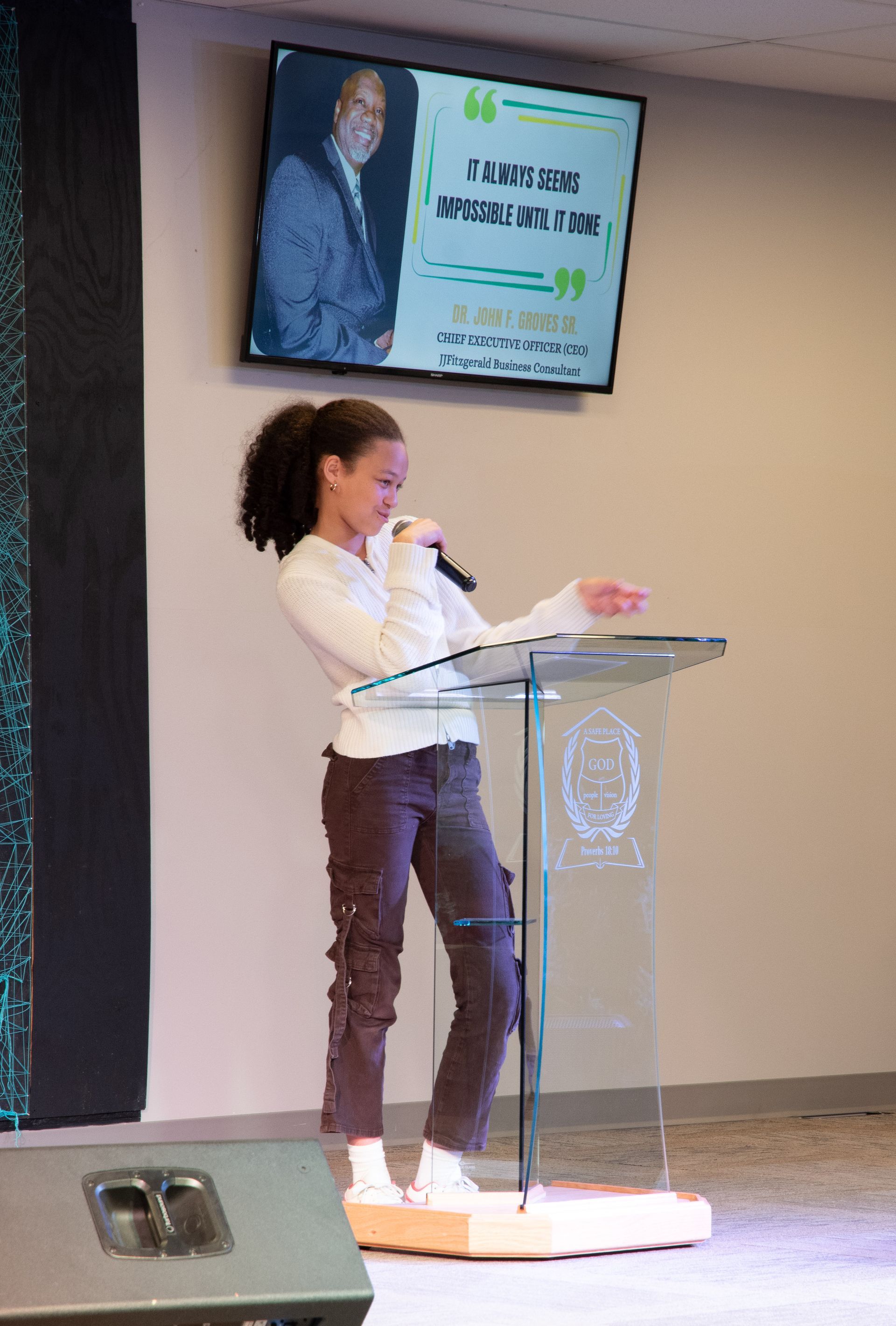 Kid Speaker at a podium presenting on stage with a slide displayed behind her.