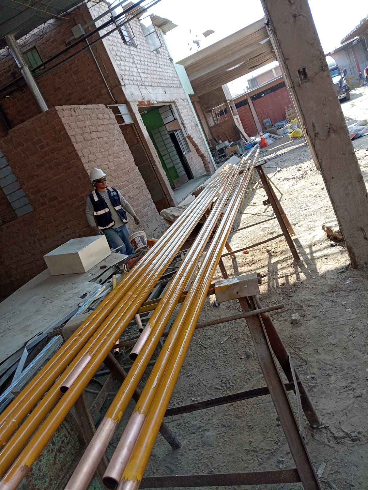 Obra: trabajador cerca de largas tuberías amarillas sobre caballetes. Edificio de ladrillo y columnas de hormigón al fondo.