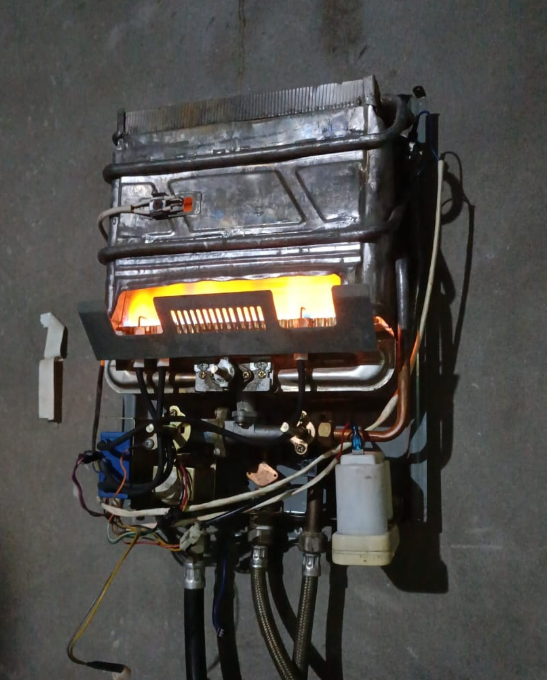 Calentador de agua a gas con llama visible. Metálico, montado en la pared.