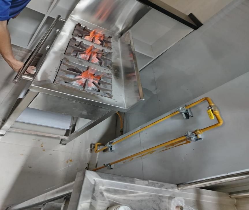 Cocina a gas comercial de acero inoxidable con llamas encendidas, montada contra una pared; líneas de gas amarillas visibles.