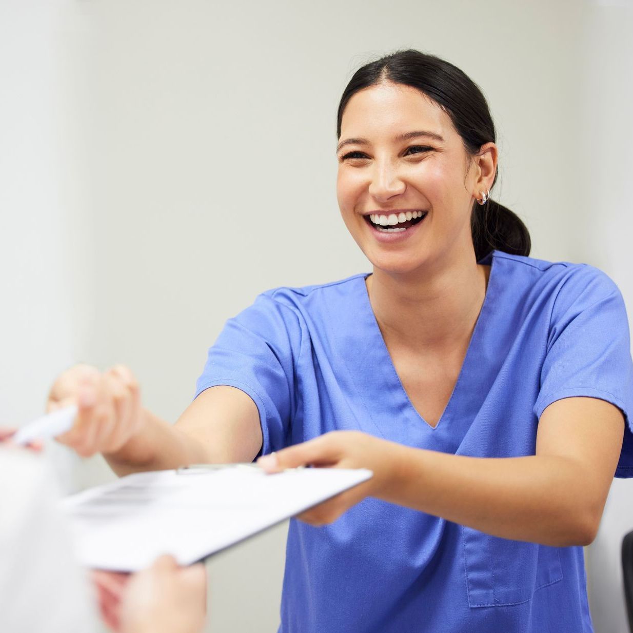 A woman in blue scrubs is smiling and holding a clipboard