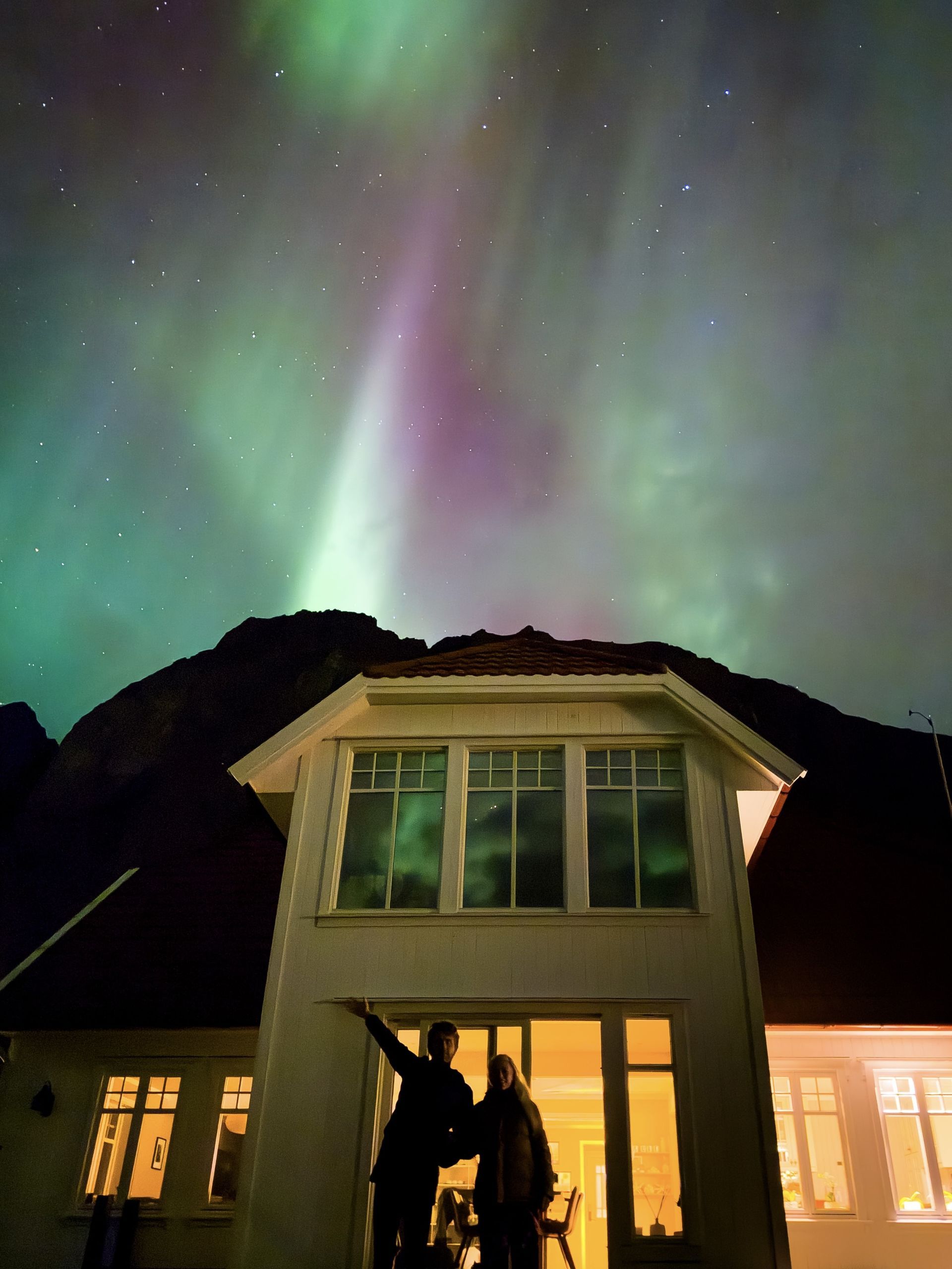 Aurora borealis over a house, two people watching, pointing upwards. Green and purple lights in the night sky.