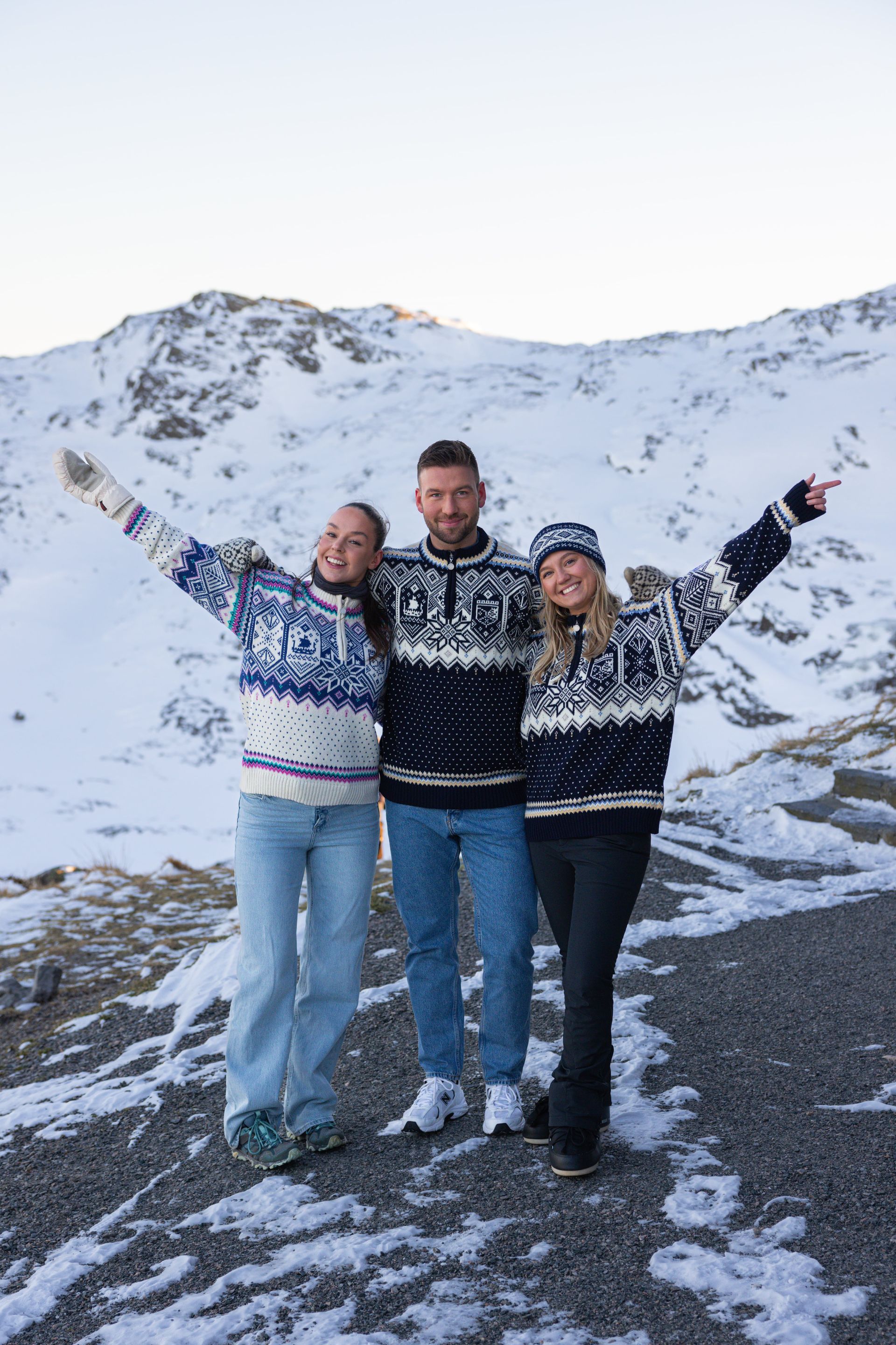 Three people in patterned sweaters pose with arms raised in snowy mountains.