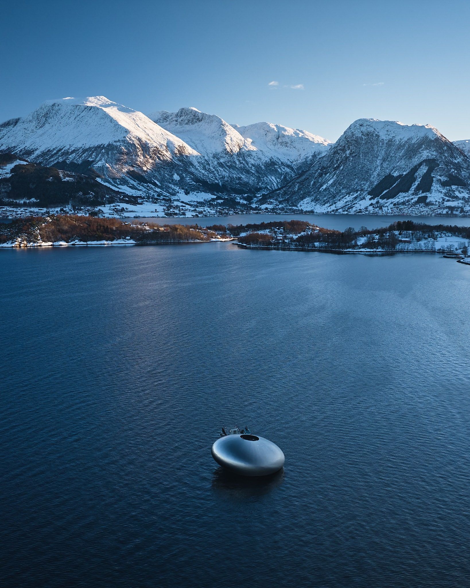 A futuristic, orb-shaped structure floats on a blue lake with snow-capped mountains in the background.